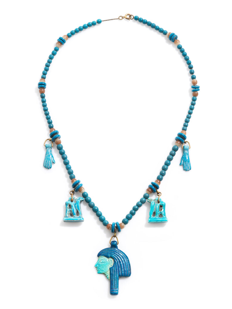 Blue hardstone necklace in Egyptian style with blue beads, with blue disc and banded agate bead stations with charms, two in the shapes of hands, 2 in the shapes of camels and a center charm of a pharaoh's head in profile. Photographed on a white background. 